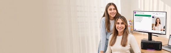 Madre e hija junto a un escritorio con un monitor LG que muestra una clase virtual. Sobre el escritorio hay un parlante portátil y materiales de estudio, en un entorno de aprendizaje en el hogar.