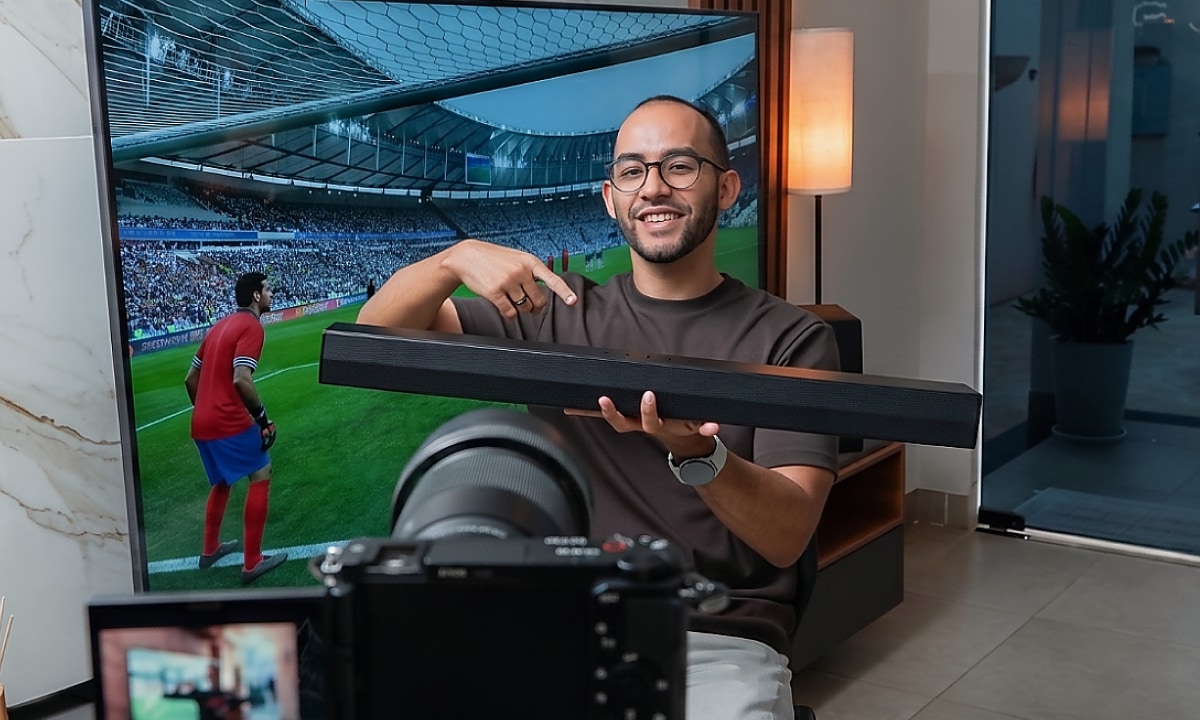Rapael sonriendo y señalando una barra de sonido que sostiene, con una TV mostrando un partido de fútbol en el fondo