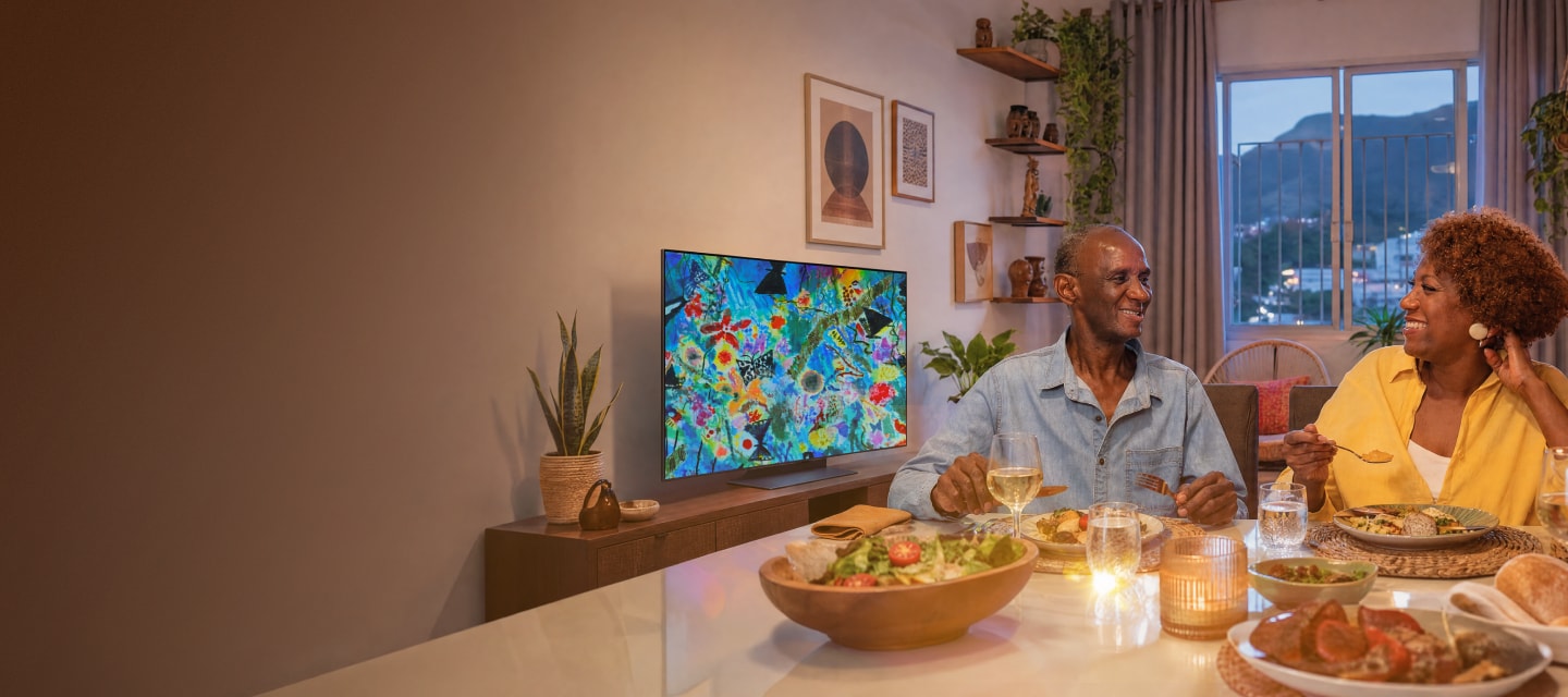 Manuel y Sandra se sonríen durante la cena mientras la TV LG OLED del fondo muestra una obra llena de color