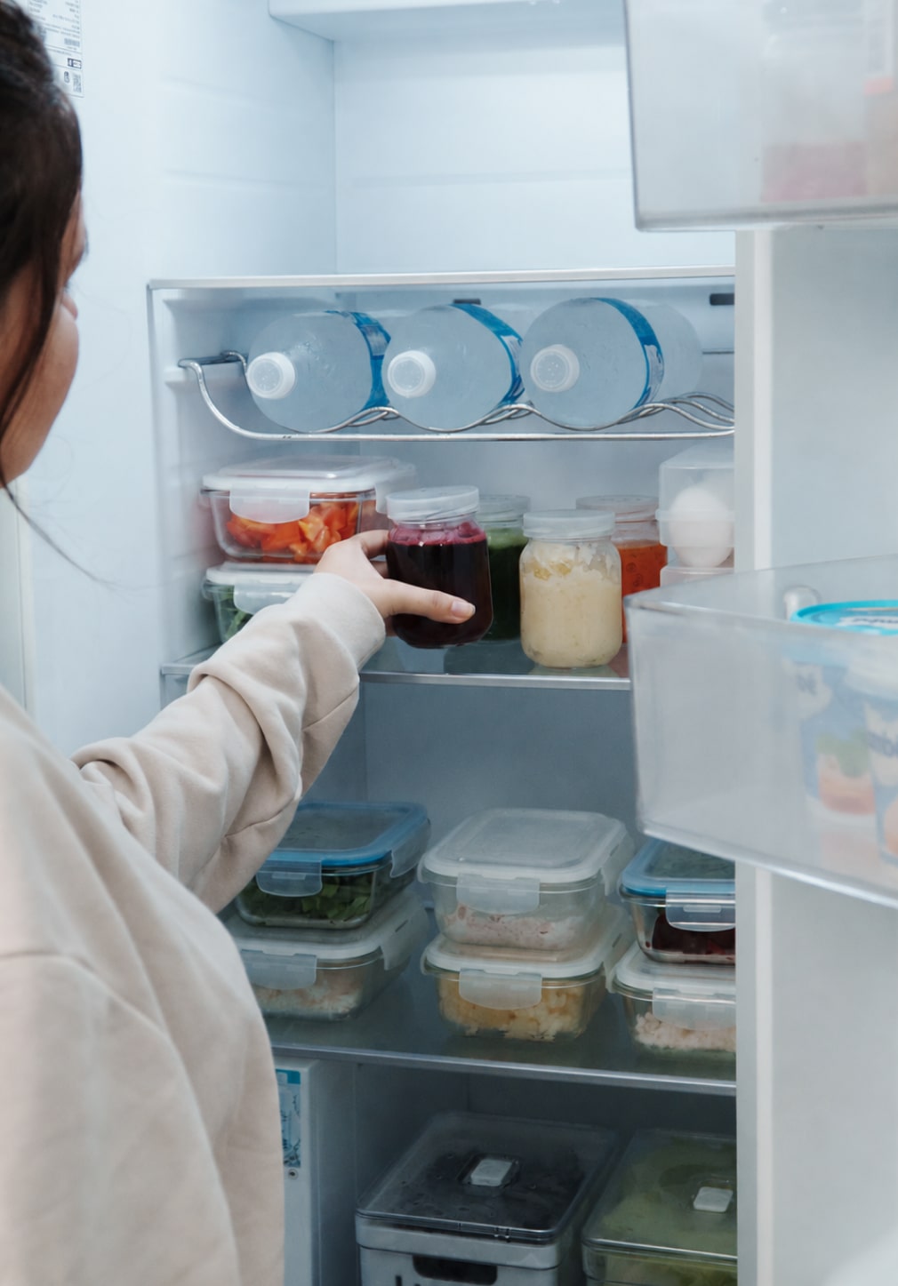 Uma mãe segura um pote dentro de uma geladeira organizada com recipientes de alimentos.