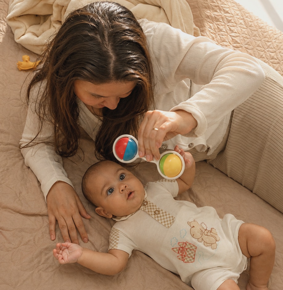 Una mamá juega con su bebé en la cama usando un sonajero de colores.