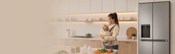 Una madre sostiene a su bebé en brazos en una cocina moderna, junto a un refrigerador LG, mientras prepara verduras frescas sobre la encimera.