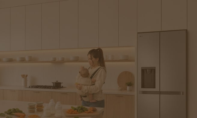 Una madre sostiene a su bebé en brazos en una cocina moderna, junto a un refrigerador LG, mientras prepara verduras frescas sobre la encimera.