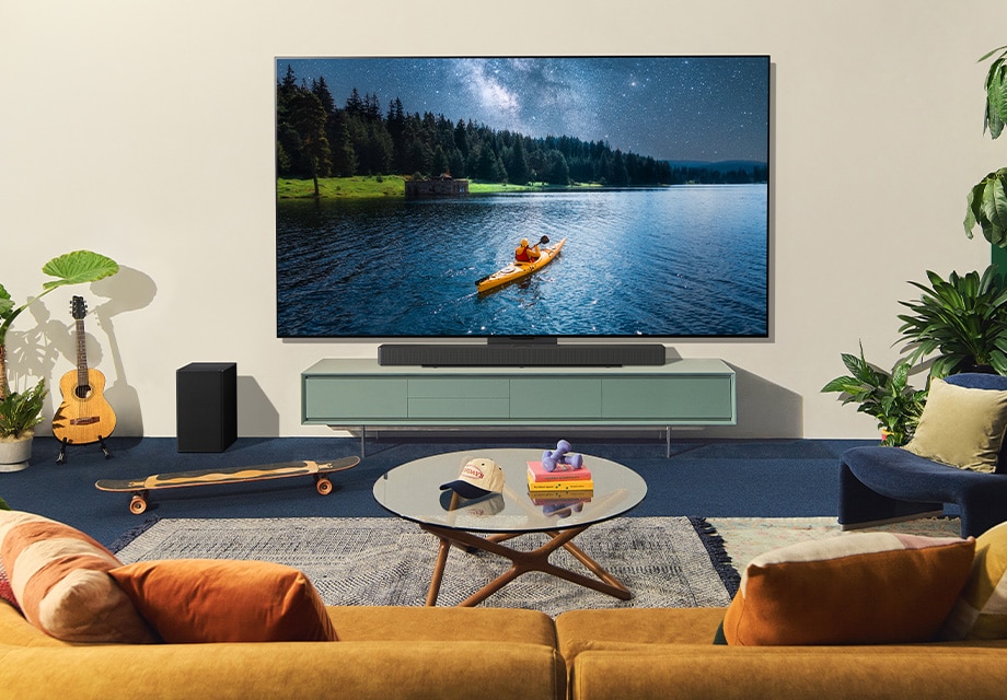 Sala de estar acogedora con dos personas viendo a un kayakista en la LG OLED TV en la pared, con plantas, guitarra, libros y muebles cálidos