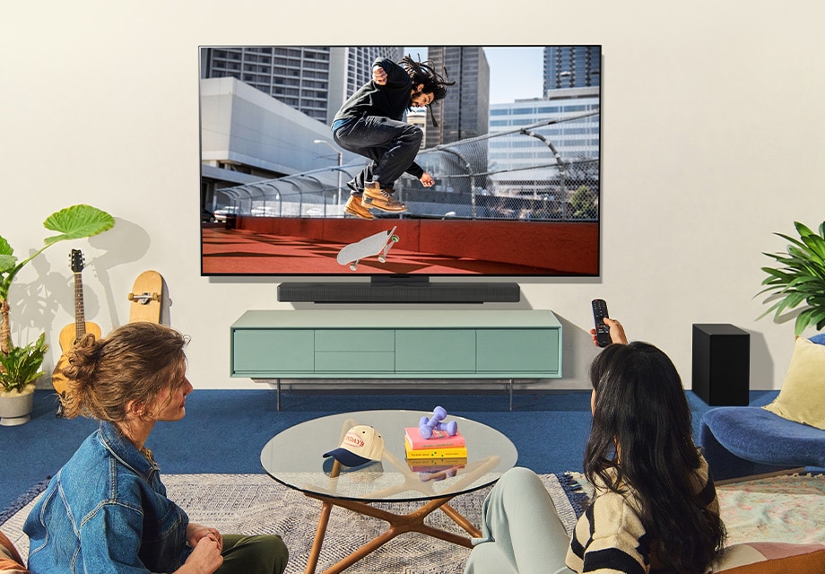 Sala de estar con dos personas viendo a un skater en la LG OLED TV montada en la pared, rodeada de plantas, libros y decoración moderna