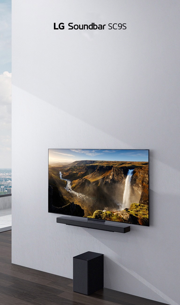 El televisor y la barra de sonido LG SC9S están colgados en una pared blanca. Debajo, hay un subwoofer inalámbrico negro en el piso. Por la ventana de la izquierda, se ve una vista de la ciudad con el cielo azul.