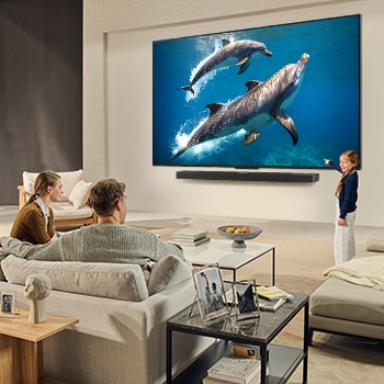 Una familia sentada en el sillón frente a la LG QNED TV montada en la pared encima de una LG Soundbar; una pequeña niña señala la pantalla que muestra dos delfines. El título habla de cómo las escenas son más emocionantes en una pantalla gigante.