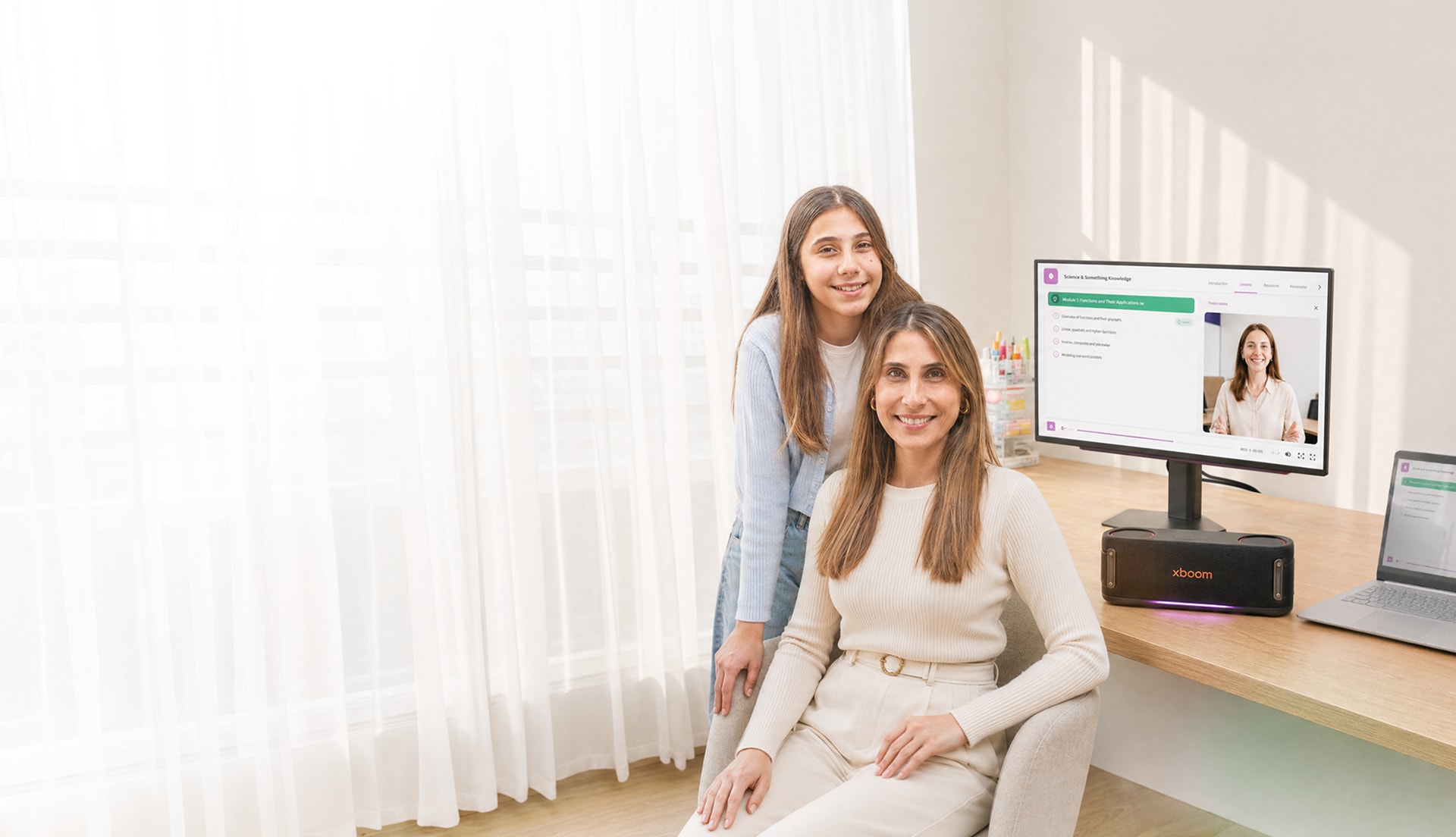 Madre sentada e hija de pie sonriendo en un escritorio iluminado con monitor LG, parlante xboom y portátil