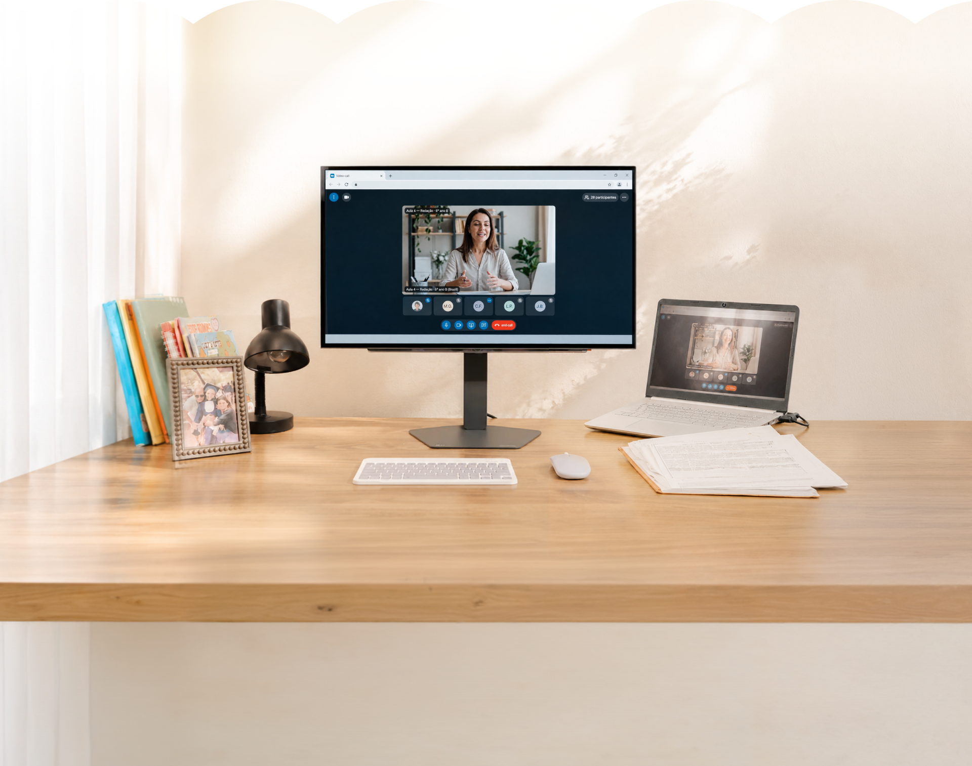Escritorio iluminado con una clase virtual por videollamada en el monitor LG, junto a un portátil, libros y lámpara