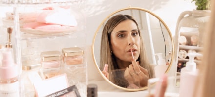 La creadora aplicándose labial frente a un espejo redondo, rodeada de productos de belleza
