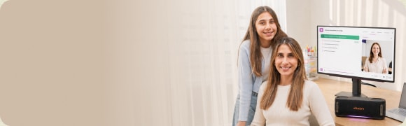 Una madre e hija sentadas frente a una computadora portátil, sonriendo. La computadora muestra una presentación de diapositivas con texto e imágenes, y hay un altavoz portátil en primer plano.