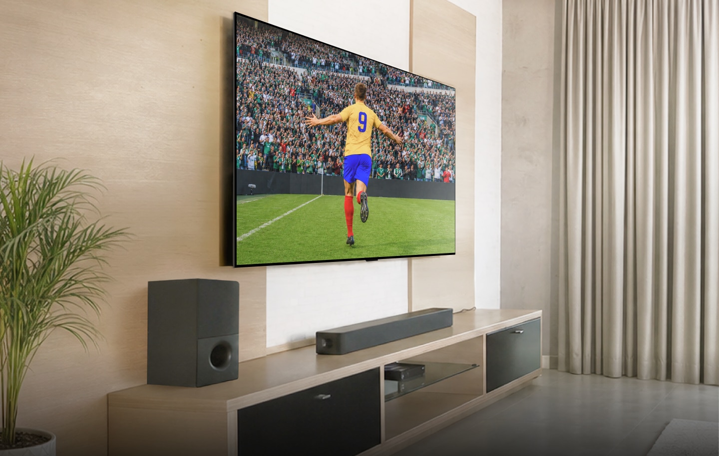 Gran televisor LG montado en la pared con una barra de sonido y un subwoofer, mostrando a un futbolista celebrando un gol.