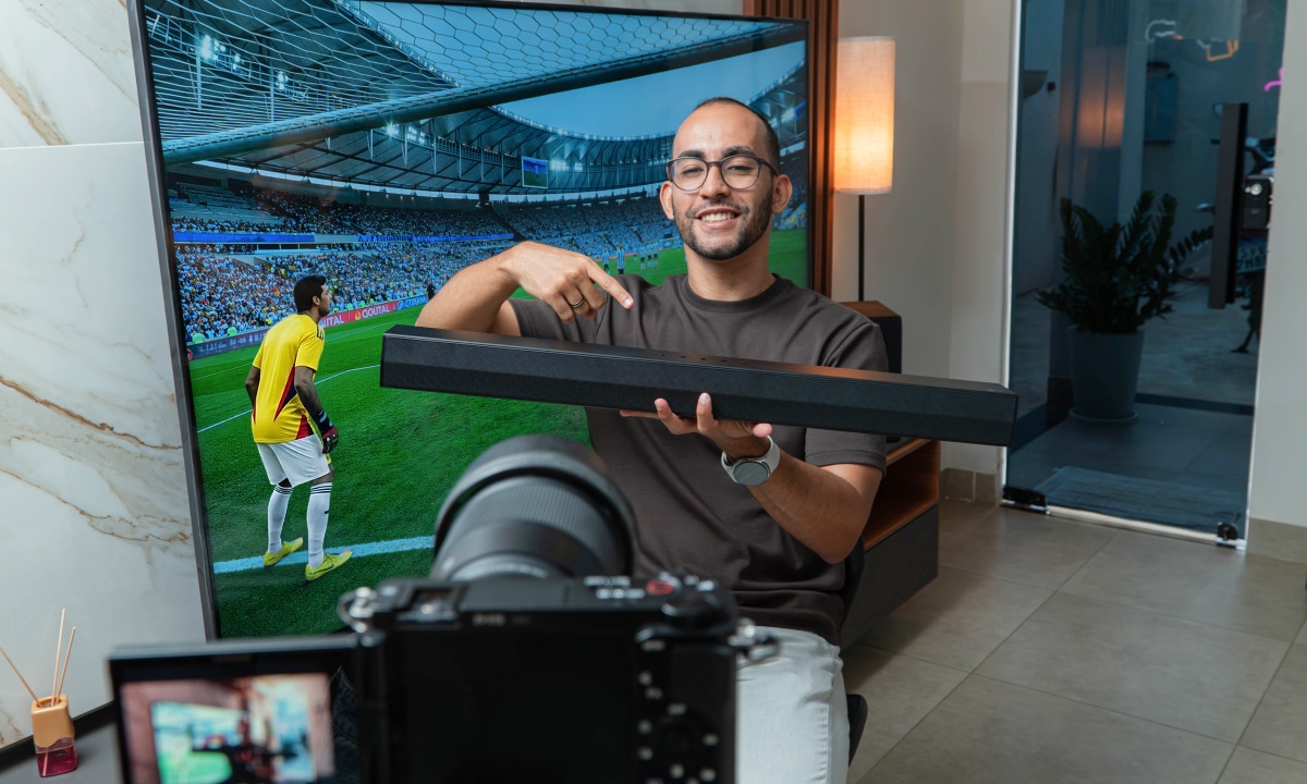 Rapael sonriendo y señalando una barra de sonido que sostiene, con una TV mostrando un partido de fútbol en el fondo