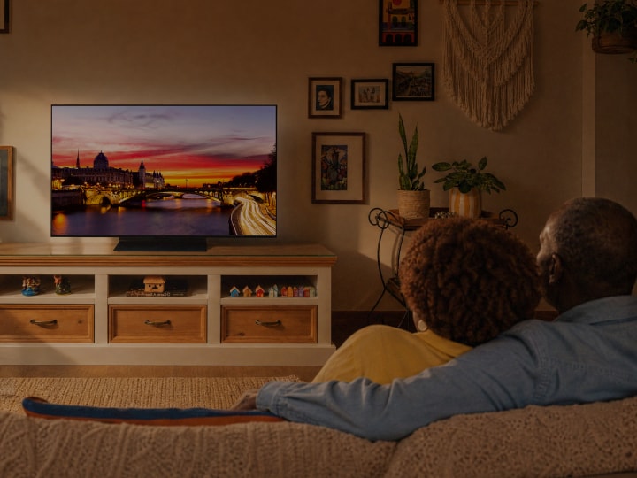 Manuel y Sandra están sentados juntos en el sofá de una sala con luz cálida, contemplando en la TV LG OLED una vista de la ciudad al atardecer.	