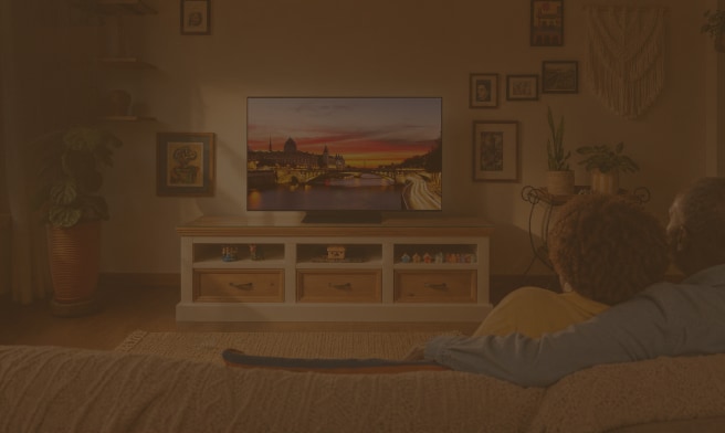 Pareja abrazada en el sofá viendo un televisor con la imagen de una ciudad al atardecer, en una sala acogedora