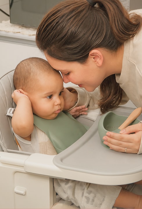 Una mamá junta su frente con la de su bebé, que está sentado en una silla alta.