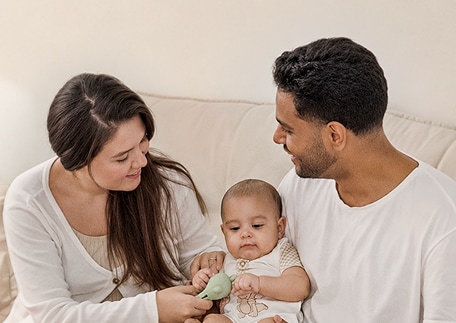 Una mamá y un papá comparten un momento con su bebé en el sofá de una sala iluminada.