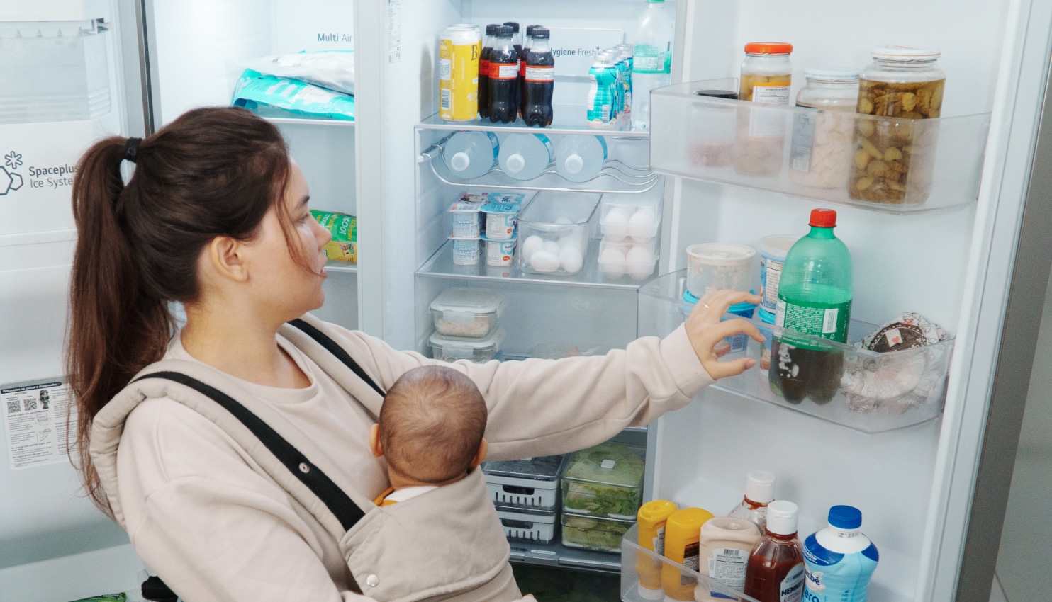 Una mamá que carga a su bebé saca una bebida de un refrigerador LG Side by Side organizado con alimentos y bebidas.