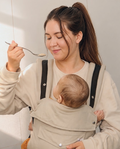 Una mamá prueba la comida con una cuchara en la cocina mientras lleva a su bebé en un portabebés.