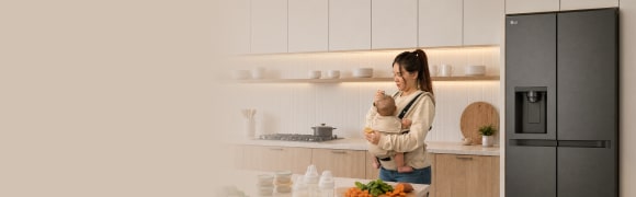 Una madre que lleva a su bebé en un portabebés prepara alimentos en una cocina moderna y luminosa, junto a un refrigerador LG.
