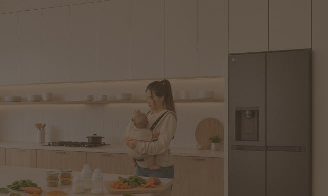 Una madre que lleva a su bebé en un portabebés prepara alimentos en una cocina moderna y luminosa, junto a un refrigerador LG.