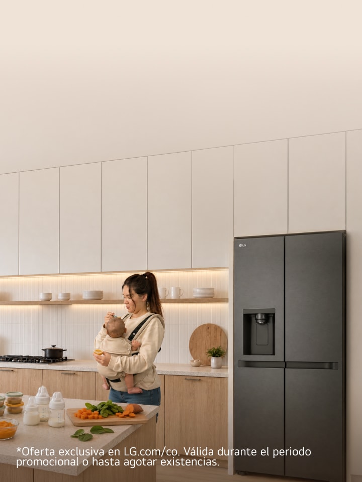 Una madre que lleva a su bebé en un portabebés prepara alimentos en una cocina moderna y luminosa, junto a un refrigerador LG.