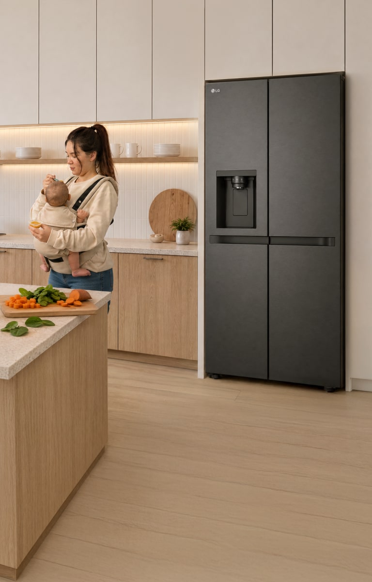 Una madre que lleva a su bebé en un portabebés prepara alimentos en una cocina moderna y luminosa, junto a un refrigerador LG.