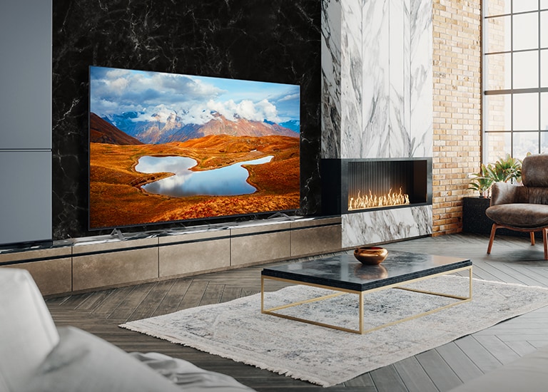 En la pared de una recepción decorada con una chimenea hay un Commercial TV grande. En la pantalla de la televisión se ven imagenes brillantes y vívidas los paisaje nocturnos de una montaña en el que se aprecia la Vía Láctea en resolución 4K