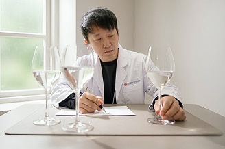  Investigador de LG Electronics con bata blanca evaluando el sabor del agua en copas de vino, en una habitación luminosa.
