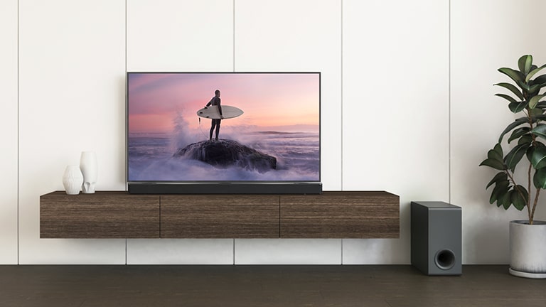 Un televisor LG y una barra de sonido LG están colocados en un estante marrón y el altavoz de subgraves está en el piso. La pantalla del televisor muestra a un surfista de pie sobre una roca.