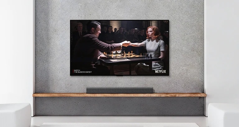 Una barra de sonido y un televisor están en un salón blanco. Una mujer y un hombre juegan al ajedrez en la pantalla del televisor.