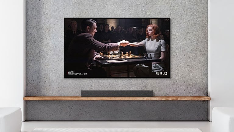 Una barra de sonido y un televisor están en un salón blanco. Una mujer y un hombre juegan al ajedrez en la pantalla del televisor.