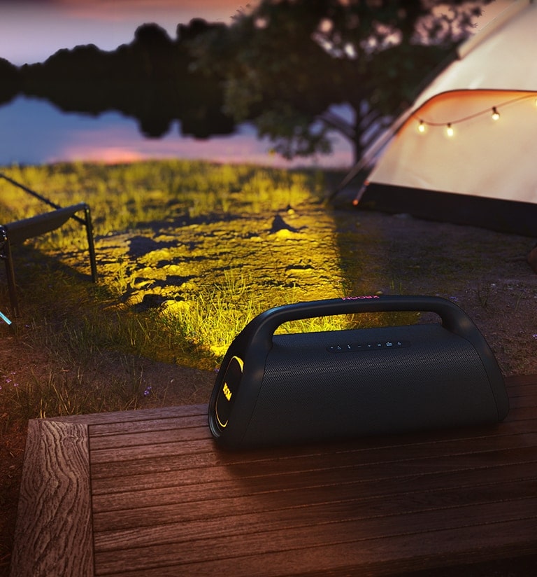 O LG XBOOM GO XG9 está colocado em um deck de madeira. Ele projeta a iluminação cênica no acampamento.