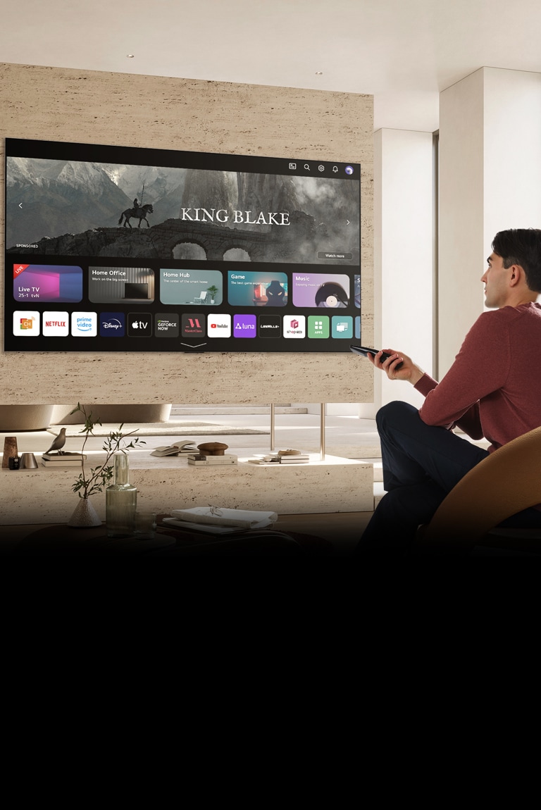 Un hombre sostiene un control remoto en su mano derecha y mira un televisor grande frente a él. La pantalla del televisor muestra la pantalla "Nuevo hogar".