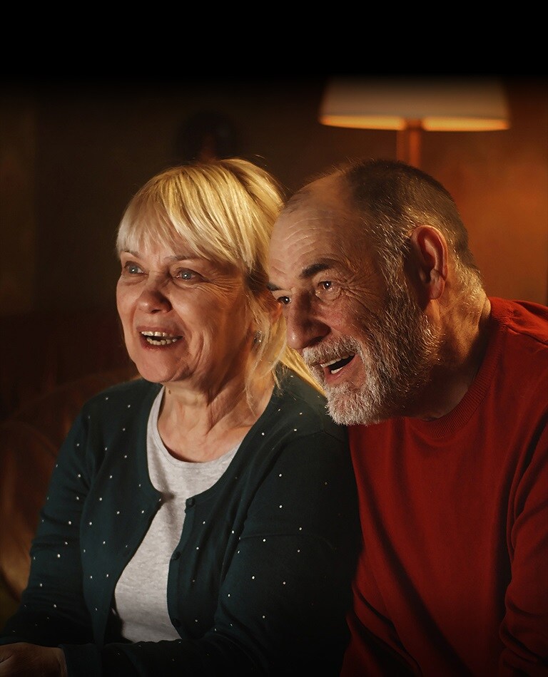Una pareja de personas mayores sentadas en un sofá, sonríen y miran la pantalla.