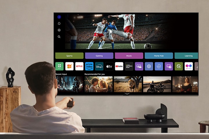 Osoba na pohovce drží v ruce dálkový ovladač. LG AI TV na stěně se systémem LG webOS na obrazovce. 