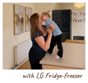 Matka drží své miminko v moderní kuchyni a stojí vedle otevřené chladničky. [with LG Fridge-freezer]