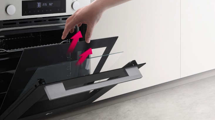 Osoba předvádí, jak v moderní kuchyni odstranit skleněný panel z vestavěných dvířek trouby.