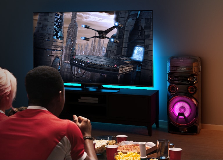 Lidé sledují TV na stěně, napravo od ní je LG XBOOM RNC7. Na televizoru běží film. 