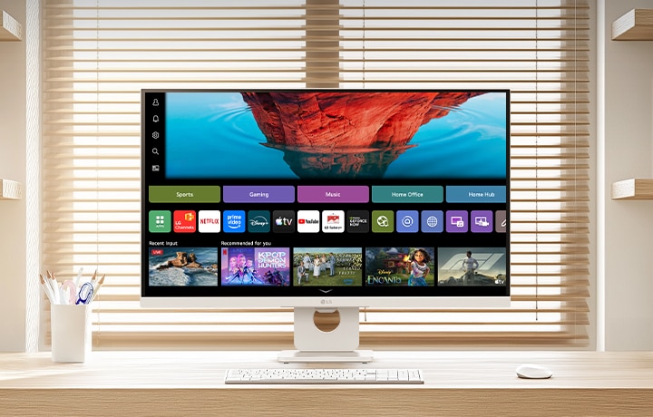 LG Smart Monitor na čistém dřevěném stole ve světlé domácí pracovně, zobrazující domovskou obrazovku webOS se streamovacími aplikacemi a dlaždicemi obsahu, doplněný klávesnicí a myší, s knihovnou a žaluziemi v pozadí, s důrazem na chytrou zábavu a produktivní využití.
