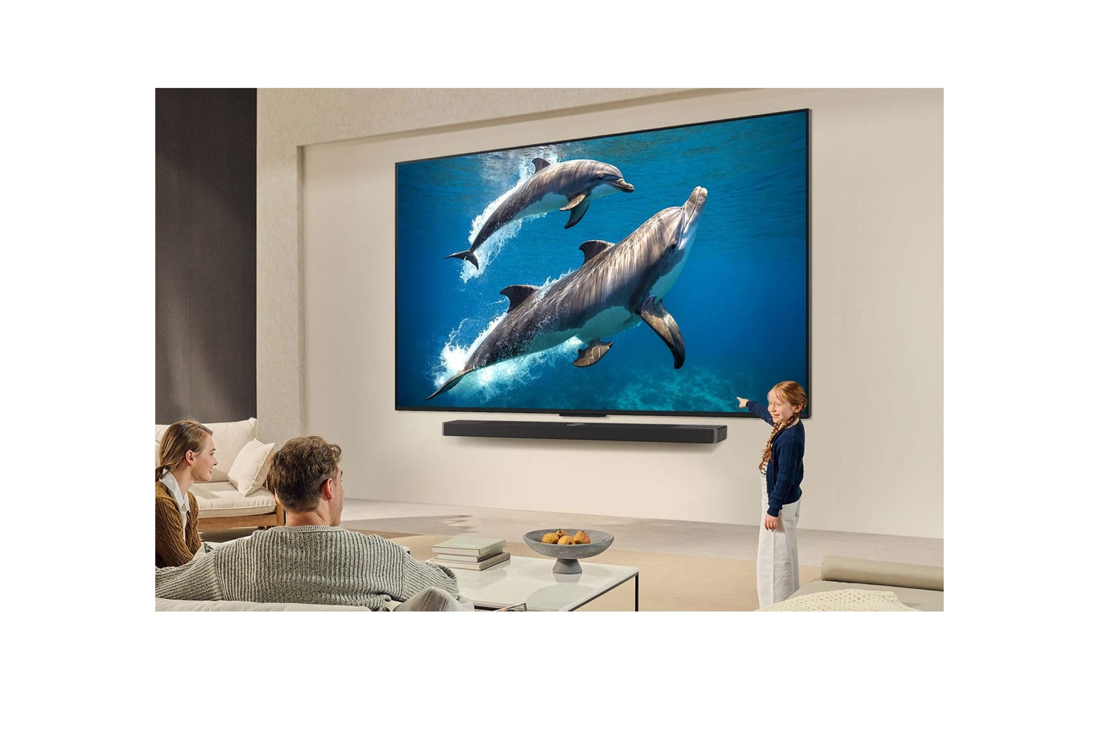 Eine Familie sitzt auf einer Couch vor einem LG QNED-Fernseher, der über einer LG Soundbar an der Wand montiert ist. Ein kleines Mädchen zeigt auf einen Bildschirm, auf dem zwei Delfine zu sehen sind. Der Titel spricht davon, dass jede Aktion auf einem großen Bildschirm noch spannender ist.