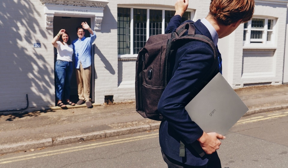 Schüler trägt Laptop und Rucksack und winkt seinen Eltern vor der Schule zum Abschied.