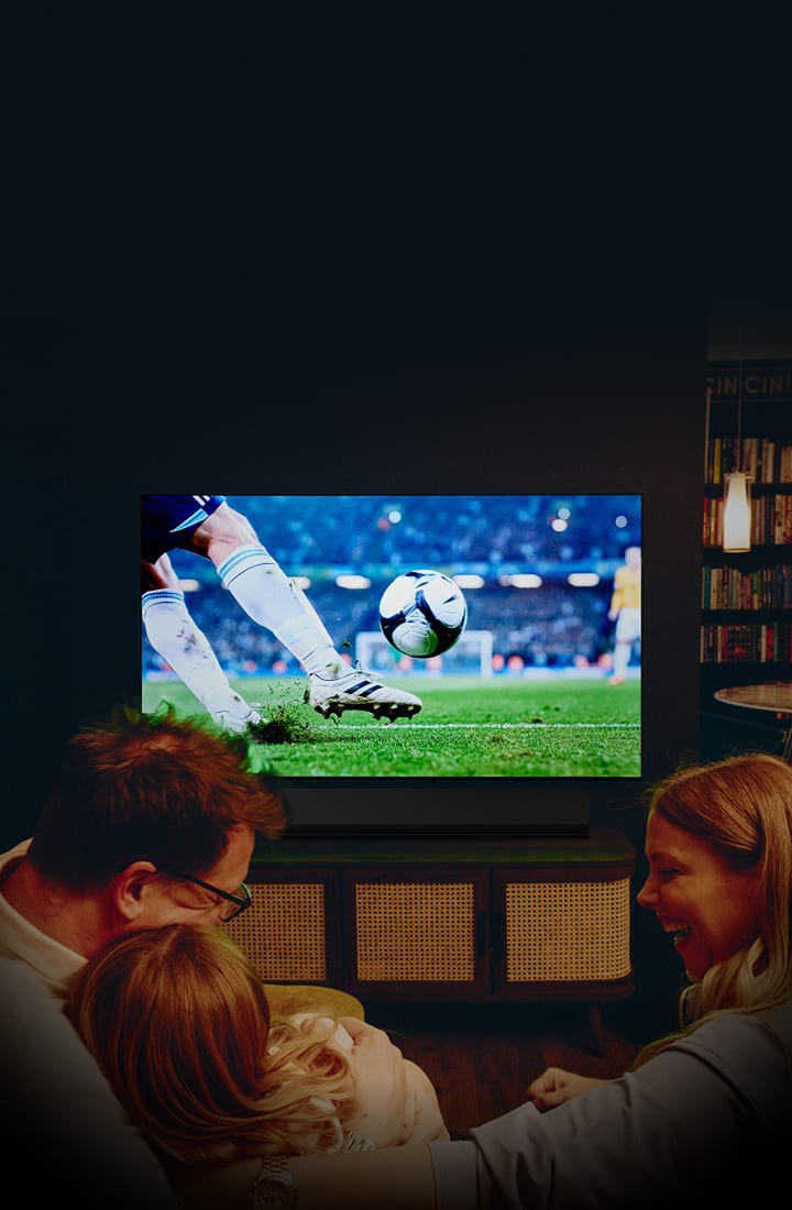 Eine Familie sitzt gemütlich im Wohnzimmer und schaut ein Fußballspiel auf einem großen wandmontierten Fernseher, auf dem eine Nahaufnahme eines Spielers zu sehen ist, der einen Ball auf einem Stadionfeld tritt, mit warmer Beleuchtung und einer kinematografischen Atmosphäre.