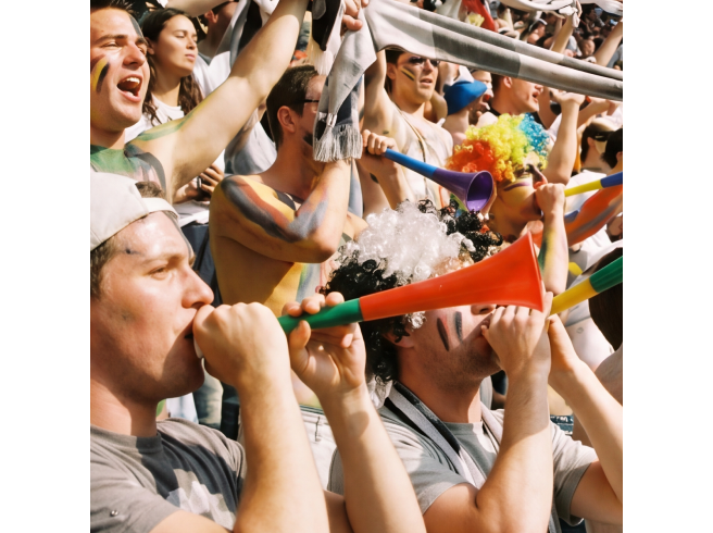 Eine Gruppe begeisterter Fans in einem Stadion jubelt und bläst bunte Vuvuzelas, trägt Teamfarben und hebt die Arme freudig in die Höhe.