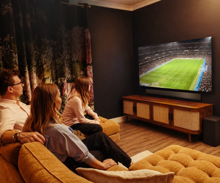 Eine dreiköpfige Familie sitzt zusammen auf einem Sofa in einem gemütlichen, gedimmt beleuchteten Wohnzimmer und schaut sich ein Fußballspiel auf einem großen LG OLED TV an. Der Raum zeichnet sich durch dunkle Wände, Blumengardinen, ein hölzernes Media-Möbel und Lautsprecher aus und schafft eine warme und intime Atmosphäre.