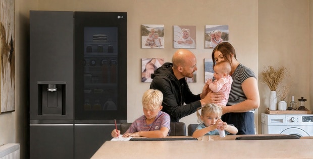 Eine Familie versammelt sich in einer Küche neben einem Edelstahl-LG-Kühlschrank – ein Junge schreibt am Esstisch, ein junges Mädchen sitzt neben ihm mit einer kleinen Flasche, und Eltern stehen zusammen und halten ein Baby.
