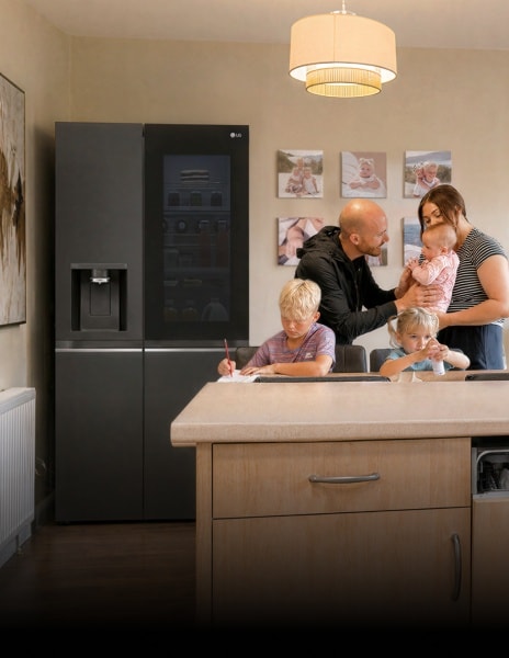 Eine Familie versammelt sich in einer Küche neben einem Edelstahl-LG-Kühlschrank – ein Junge schreibt am Esstisch, ein junges Mädchen sitzt neben ihm mit einer kleinen Flasche, und Eltern stehen zusammen und halten ein Baby.