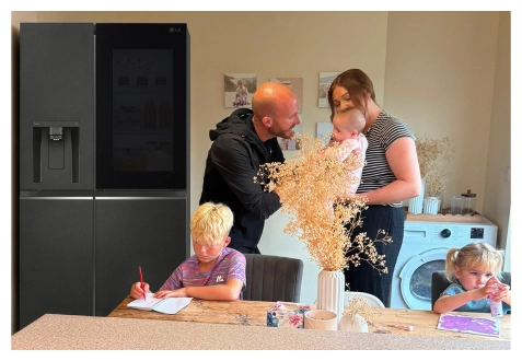 Eine Familie versammelt sich in der Küche neben einem Edelstahl-Kühlschrank – ein Junge zeichnet am Tisch, ein Mädchen malt an der Arbeitsfläche, und die Eltern stehen zusammen und halten ein Baby.