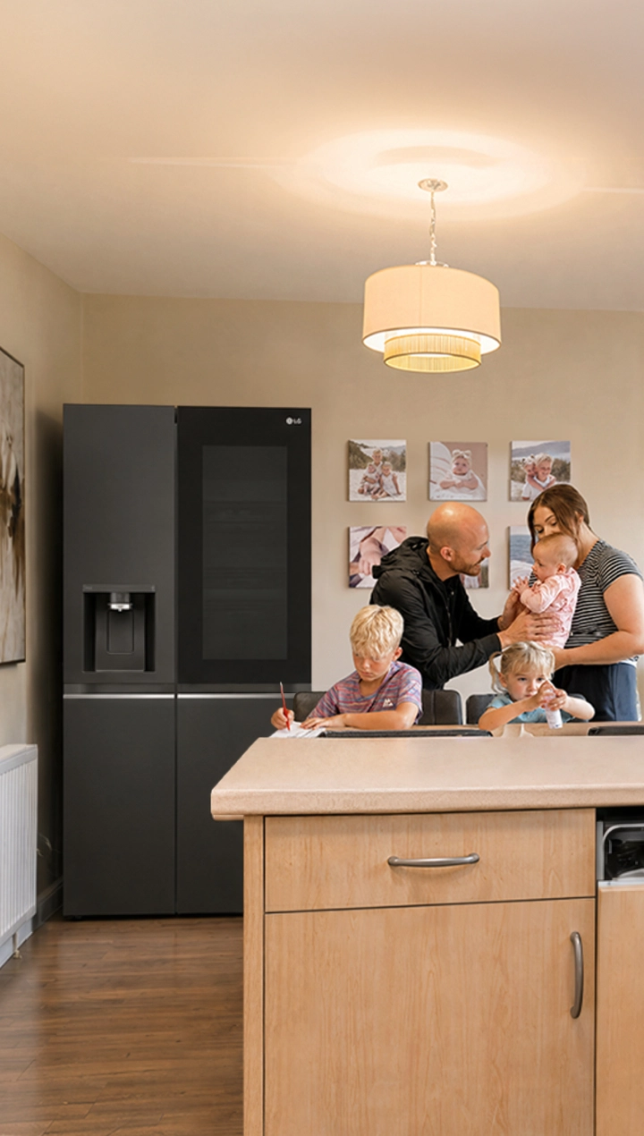Eine Familie mit drei Kindern versammelt sich in einer warmen, modernen Küche, wobei sich die Eltern um die Kinder an einer großen Kücheninsel kümmern. Ein Kühlschrank, eine Spülmaschine, ein Ofen und Küchenschränke sind im Hintergrund sichtbar.