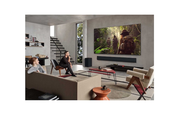 Weite Ansicht eines Wohnzimmers in einem hochwertigen Haus. Ein Mann sieht sich einen Film auf seinem an der Wand montierten LG OLED-Fernseher an. Das Design des Fernsehers fügt sich elegant in die Wand und die Inneneinrichtung ein.